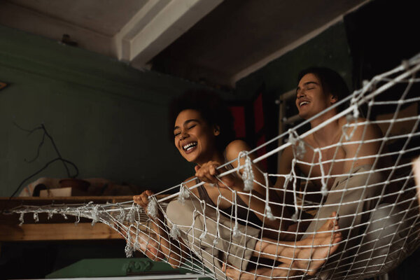 excited african american woman laughing in hammock near smiling shirtless boyfriend 