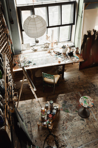 spacious workshop with large window and art tools on windowsill near chair and easel 