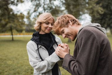 caring young man in coat warming up hands of happy girlfriend in autumnal park  clipart