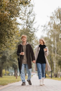 Paltolu mutlu bir çiftin el ele tutuşması ve sonbahar parkında kağıt bardaklarla yürümesi. 