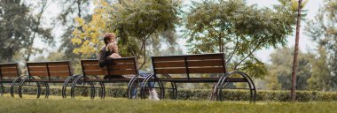cheerful young man and woman hugging and sitting on wooden bench in green park, banner clipart