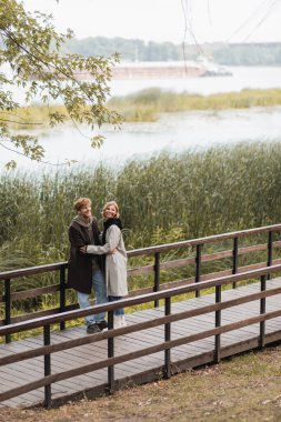 Uzun boylu kızıl saçlı adam ve ceketli mutlu kadın parkta, gölün kenarında köprüde kucaklaşırken gülümsüyor. 