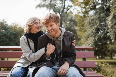 cheerful and blonde woman embracing redhead boyfriend while sitting on bench in park clipart
