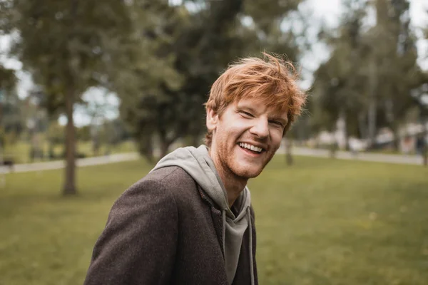cheerful and redhead man in autumnal outfit smiling in park