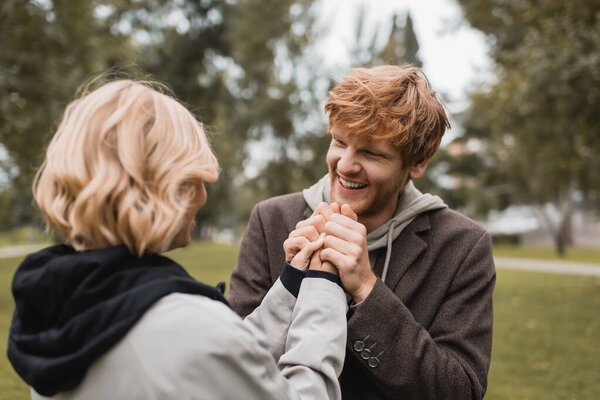 caring young man in coat warming up hands of blonde girlfriend in autumnal park  