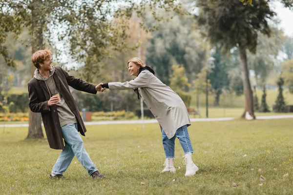 コート姿の若い男が紙コップを持って秋の公園で彼女を引っ張っ — ストック写真
