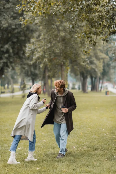コートに身を包んだ喜びの男が紙コップを手に秋の公園で喜ぶ彼女の手 — ストック写真