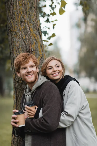 Happy Blonde Woman Holding Paper Cup Hugging Boyfriend Tree Trunk — Stock Photo, Image