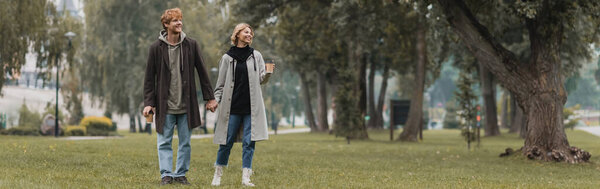 full length of happy redhead man and cheerful woman holding coffee to go while walking in park, banner