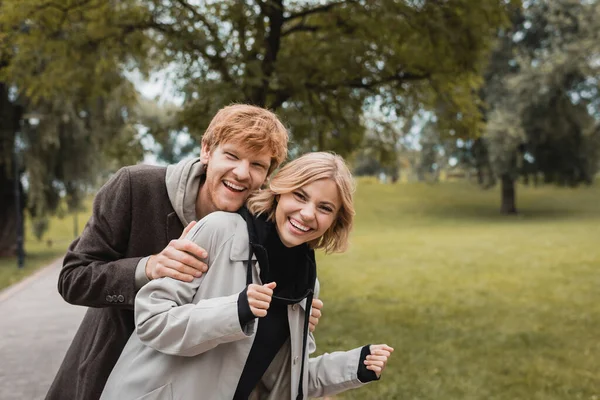 快乐的男人一边抱着快乐的年轻女人一边看着公园里的相机 — 图库照片