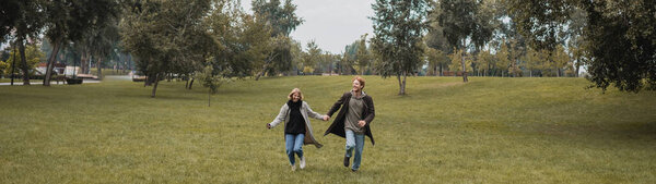 full length of happy man and pleased woman in coats holding hands while running on grass, banner