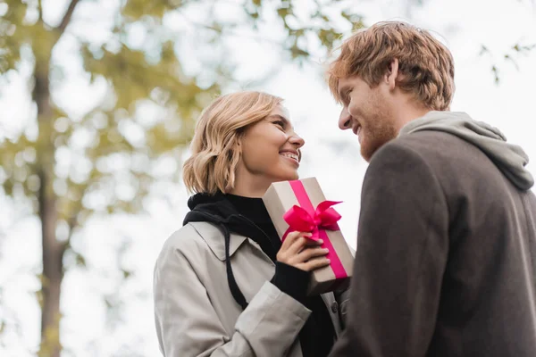 low angle view of happy woman holding wrapped gift box with pink ribbon and looking at boyfriend in park