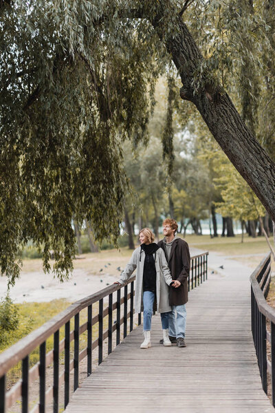 full length of redhead man and blonde woman in coat smiling while holding hands on bridge near pond in park 