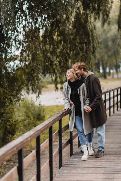 full length of redhead man and blonde woman in coat smiling while having date in park 