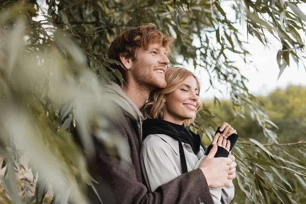 Šťastný Muž Objímání Veselá Mladá Žena Blízkosti Zelené Listy Rozmazané — Stock fotografie