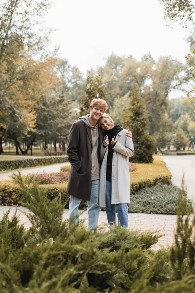 Fröhlicher Junger Mann Mantel Umarmt Blonde Freundin Während Park Pflanzen — Stockfoto