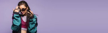 Woman in vintage clothes and sunglasses looking at camera isolated on purple, banner  clipart