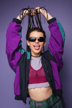 young woman in vintage jacket and sunglasses holding braids and sticking out tongue isolated on purple clipart