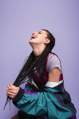 excited woman in nineties style outfit holding braided dreadlocks and laughing isolated on purple clipart