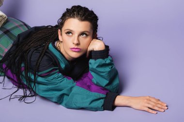 Eski moda ceket içinde uzanmış, mor arka planda gözlerini kaçıran hayalperest bir kadın.