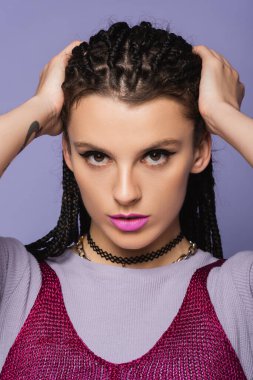 portrait of pretty woman in makeup touching braided hair while looking at camera isolated on purple clipart