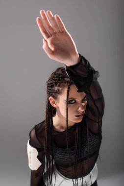 high angle view of woman with braids and futuristic style makeup and clothes posing with raised hand isolated on grey clipart