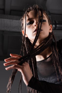 low angle view of stylish woman with braided dreadlocks looking at camera on grey background clipart