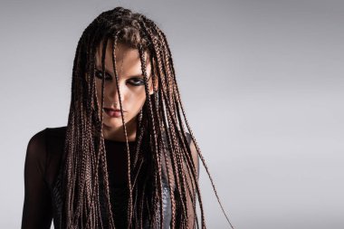 portrait of young woman with stylish makeup and braided dreadlocks looking at camera isolated on grey clipart
