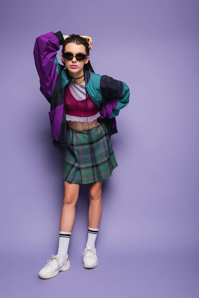 Fashionable woman in sports jacket and sunglasses posing on purple background 