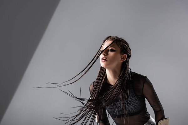 молодая брюнетка с косичками и модным макияжем позирует на сером фоне