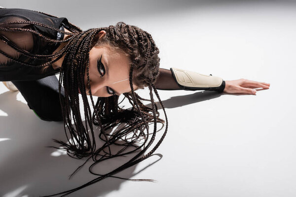 чувственная брюнетка женщина с футуристическим макияжем и косы, позирующие на белой поверхности и сером фоне