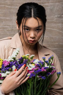 Portrait of brunette asian woman in trench coat holding flowers on abstract brown background  clipart