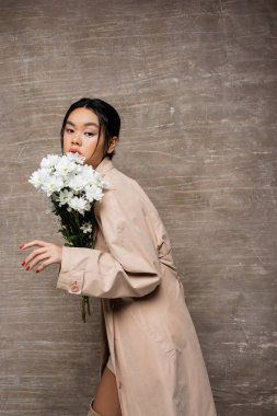 Pretty asian woman in trench coat posing with white chrysanthemums on abstract brown background  clipart