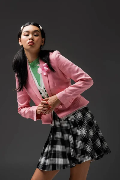 Pretty asian woman in skirt and pink jacket looking at camera isolated on grey