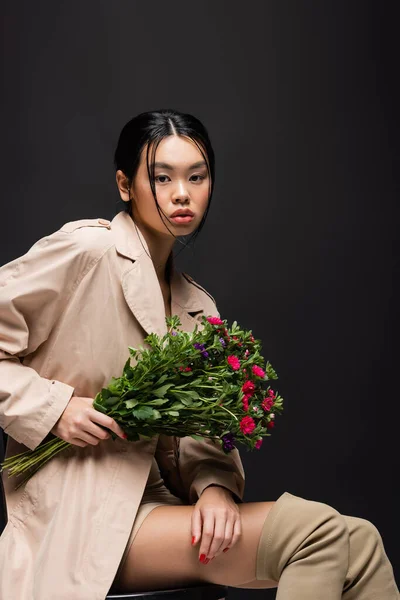 Pretty asian woman in boots and trench coat holding colorful bouquet isolated on black