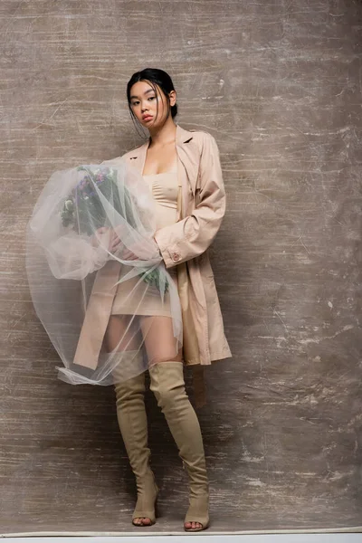 Stylish asian model in dress and trench coat holding flowers under veil on abstract brown background