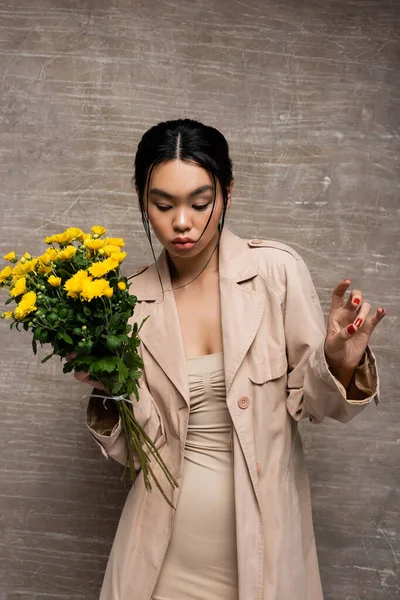 Young asian woman in trench coat and dress holding yellow flowers on abstract brown background