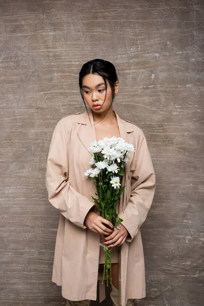 Trençkotlu Asyalı Kadın Soyut Kahverengi Arka Planda Beyaz Kasımpatı Tutuyor — Stok fotoğraf