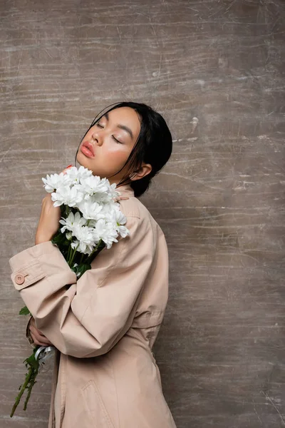 Модная Азиатка Плаще Держит Хризантемы Закрывает Глаза Абстрактном Коричневом Фоне — стоковое фото