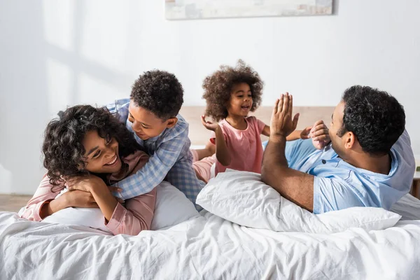 Щасливий афроамериканський хлопчик обіймає матір біля тата і сестру, граючи в торт в ліжку — стокове фото