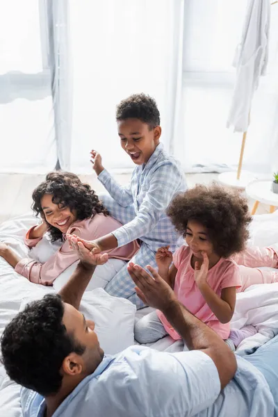 Afro-americano homem jogar Patty bolo jogo com crianças na cama perto alegre esposa — Fotografia de Stock