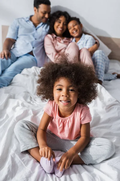 Gioiosa ragazza afro-americana sorridente alla macchina fotografica vicino a genitori e fratello sfocati in camera da letto — Foto stock