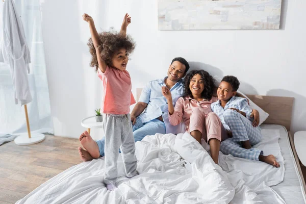 Aufgeregtes afrikanisch-amerikanisches Mädchen, das mit erhobenen Händen neben lächelnden Eltern und Bruder auf dem Bett steht — Stockfoto