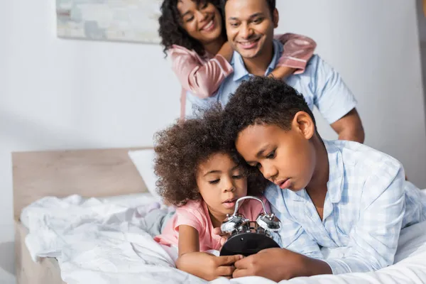 Crianças afro-americanas olhando para despertador perto de pais desfocados sorrindo no quarto — Fotografia de Stock