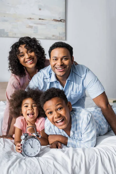 Cheerful african kids with alarm clock and smiling parents looking at camera in bedroom — Stock Photo