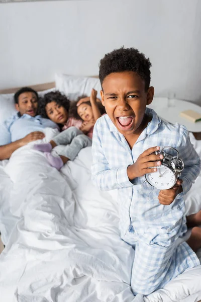 Emocionado afroamericano chico con despertador gritando cerca borrosa familia acostado en la cama - foto de stock