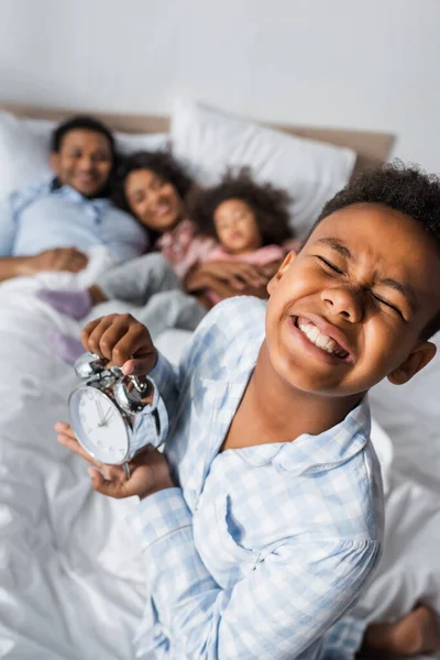 Aufgeregter afrikanisch-amerikanischer Junge mit geschlossenen Augen hält Wecker in der Nähe verschwommener Familie im Bett — Stockfoto