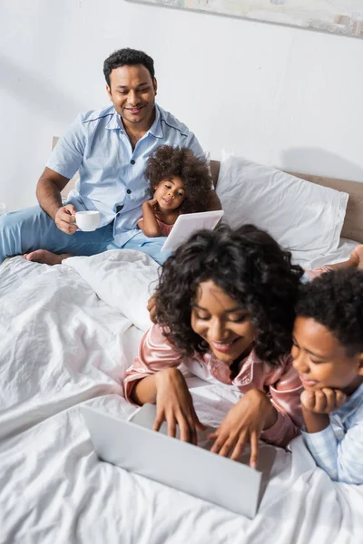 Feliz afroamericana mujer escribiendo en el ordenador portátil cerca de hijo y borrosa marido con hija - foto de stock