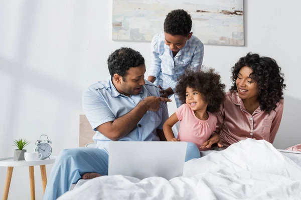 Африканский американец с ноутбуком указывает на дочь рядом с сыном и женой в спальне — стоковое фото