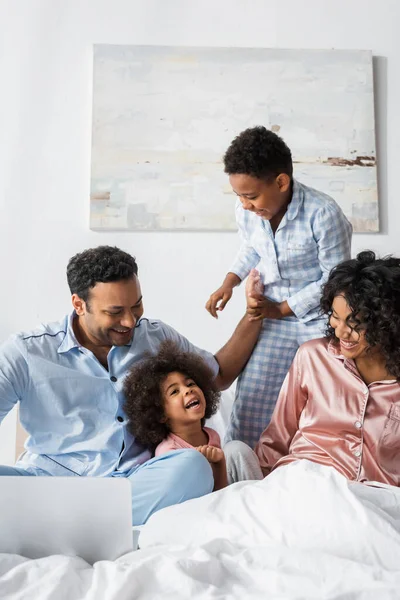 Africano americano uomo sorridente vicino laptop e felice famiglia sul letto — Foto stock